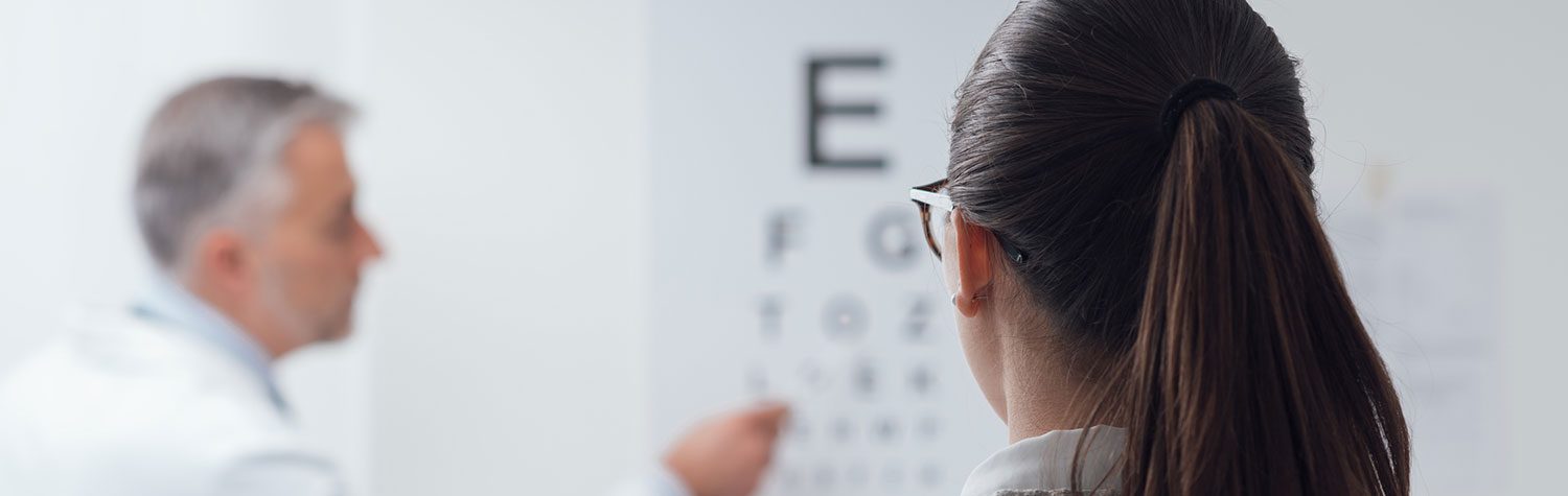 Facing away from the camera, a woman looks at an eye chart during an eye exam