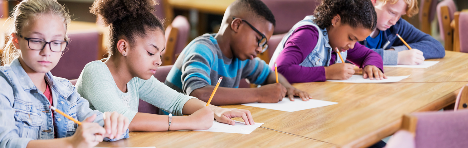 Five children writing with pencil and paper while sitting in a row at a long wooden table