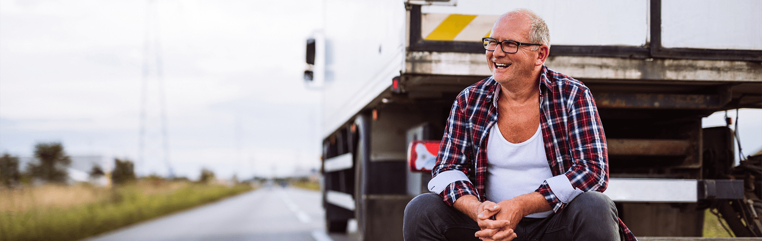 Smiling middle aged man in plaid shirt and tank top sits on the road behind his box truck