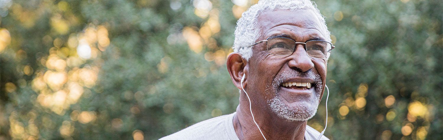 An older man smiling and wearing headphones in an outdoor setting.