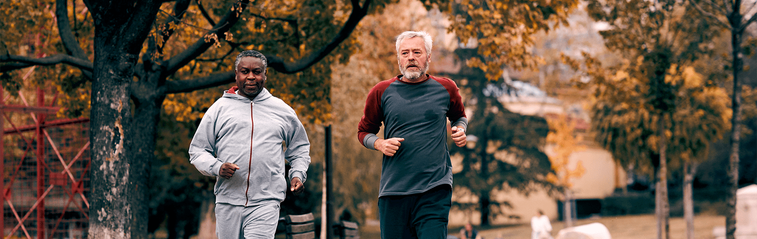 Two older men running together on an outdoor, tree-lined trail.