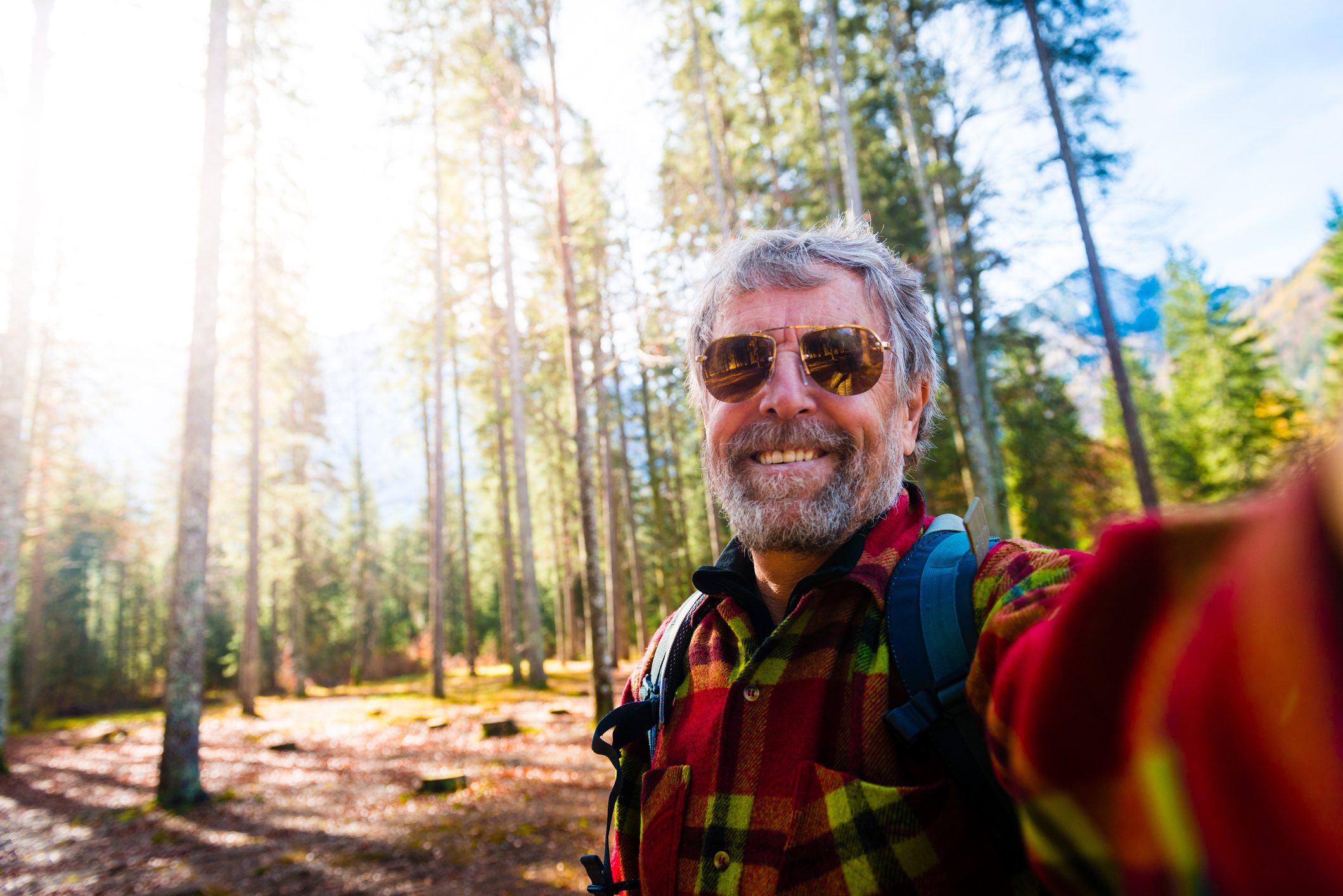 An older man in a flannel and sunglasses taking a selfie on a sunny day in the forest