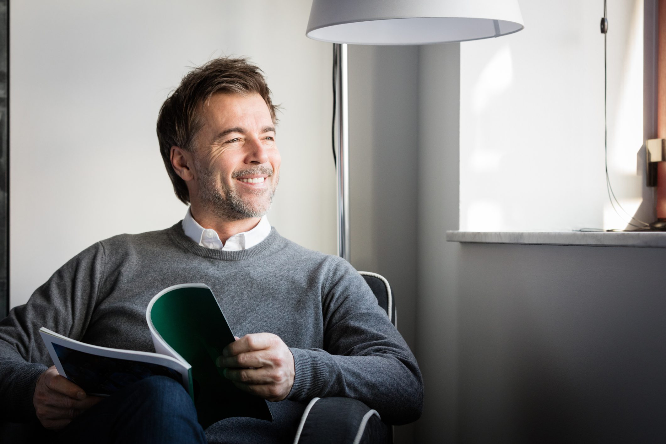 A brown-haired man sitting on a chair with a book and smiling out the window