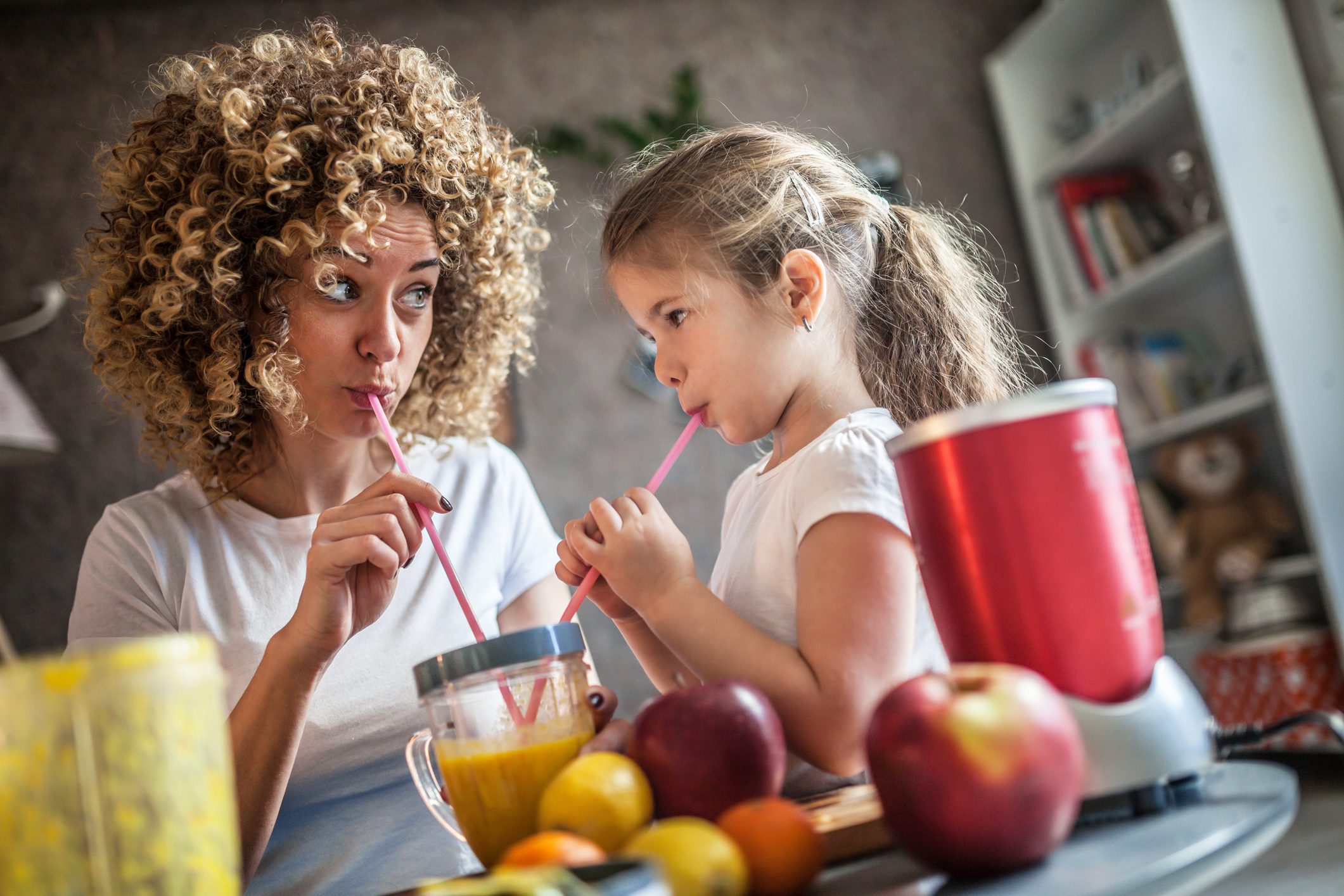 A mother and daughter drink a smoothie from long pink straws in a kitchen