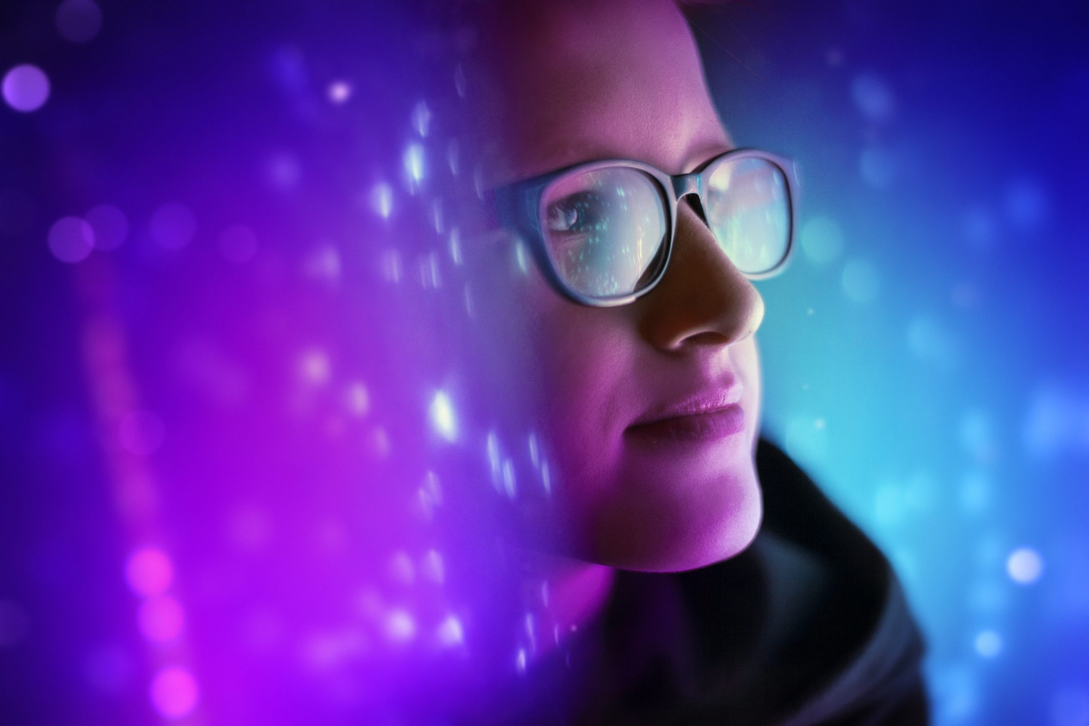 A woman with glasses stands in a dark room lit up with bright blue and purple colors.