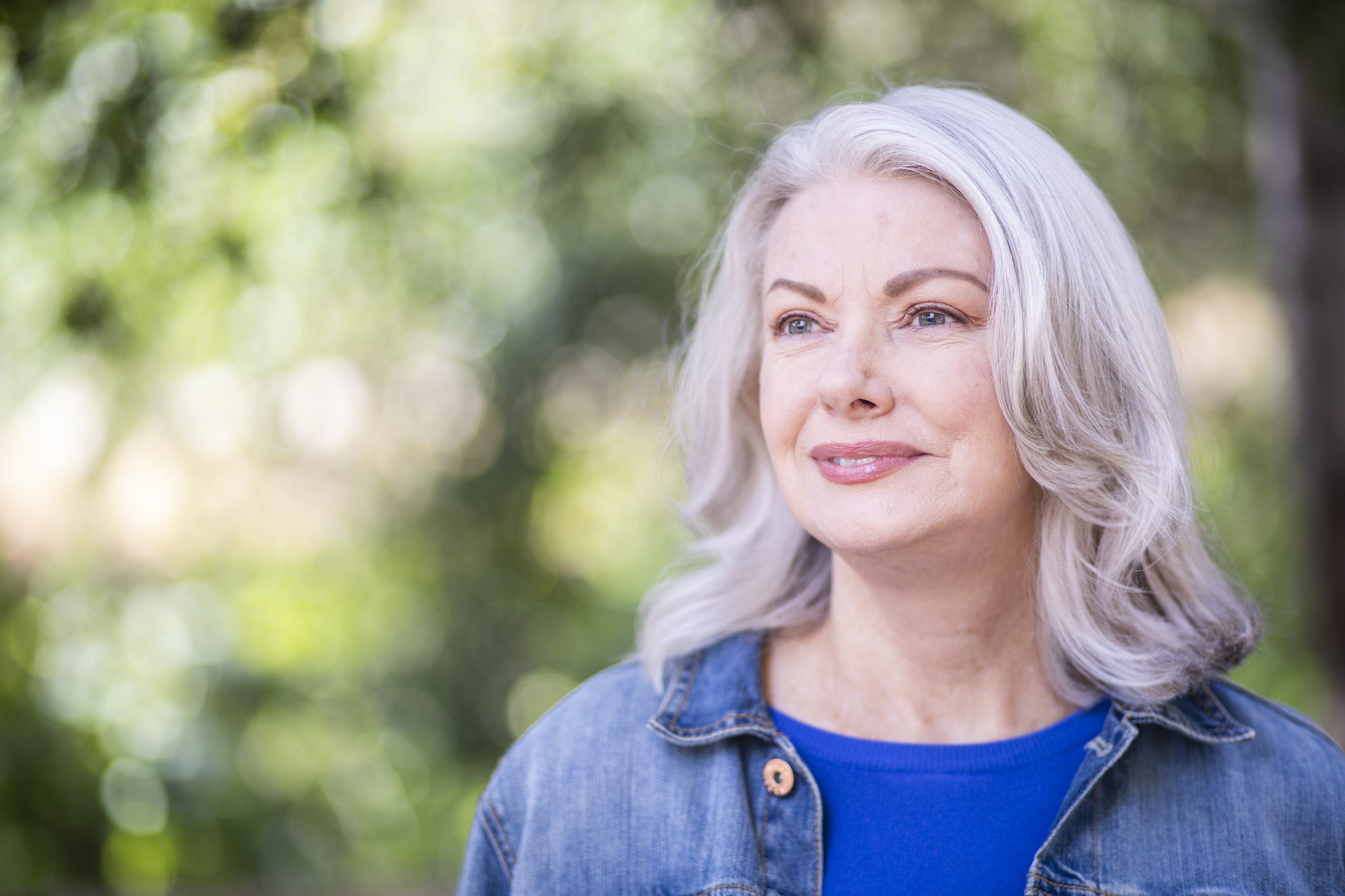 Older woman with shoulder-length white hair and a denim jacket smirks at something off camera while standing in front of a blurred outdoor background