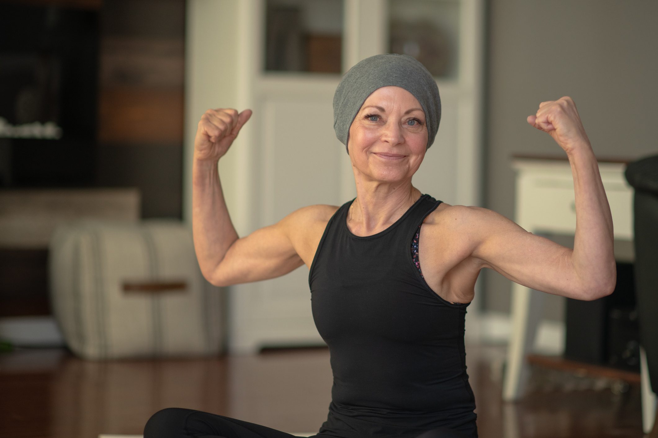 An older woman wearing a tight beanie sits crosslegged on the floor and flexes her muscles