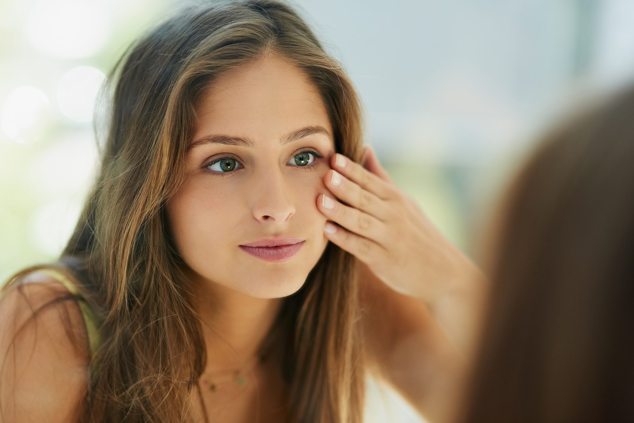 A young woman touches her face while looking in a mirror
