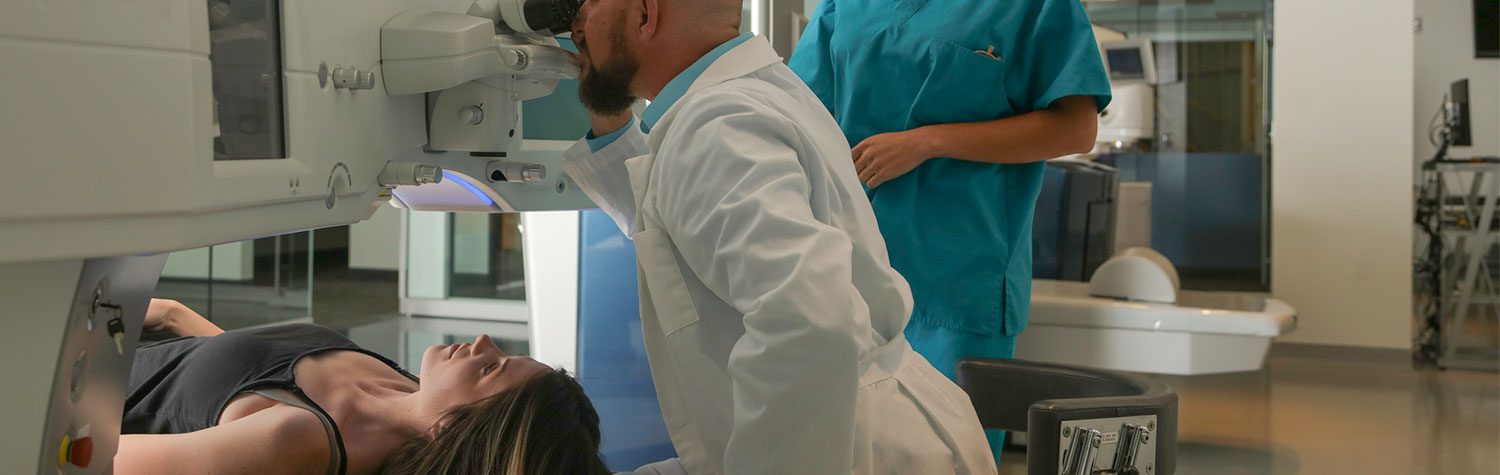 A woman lays on her back in a medical machine as a male doctor looks into the machine and a nurse watches nearby