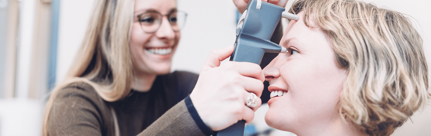 Blonde woman in glasses gives an eye pressure test to a young blonde female patient.
