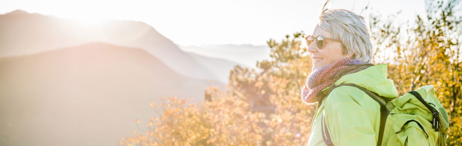 Older woman in a green jacket and backpack hiking in sunglasses with mountains in the background.