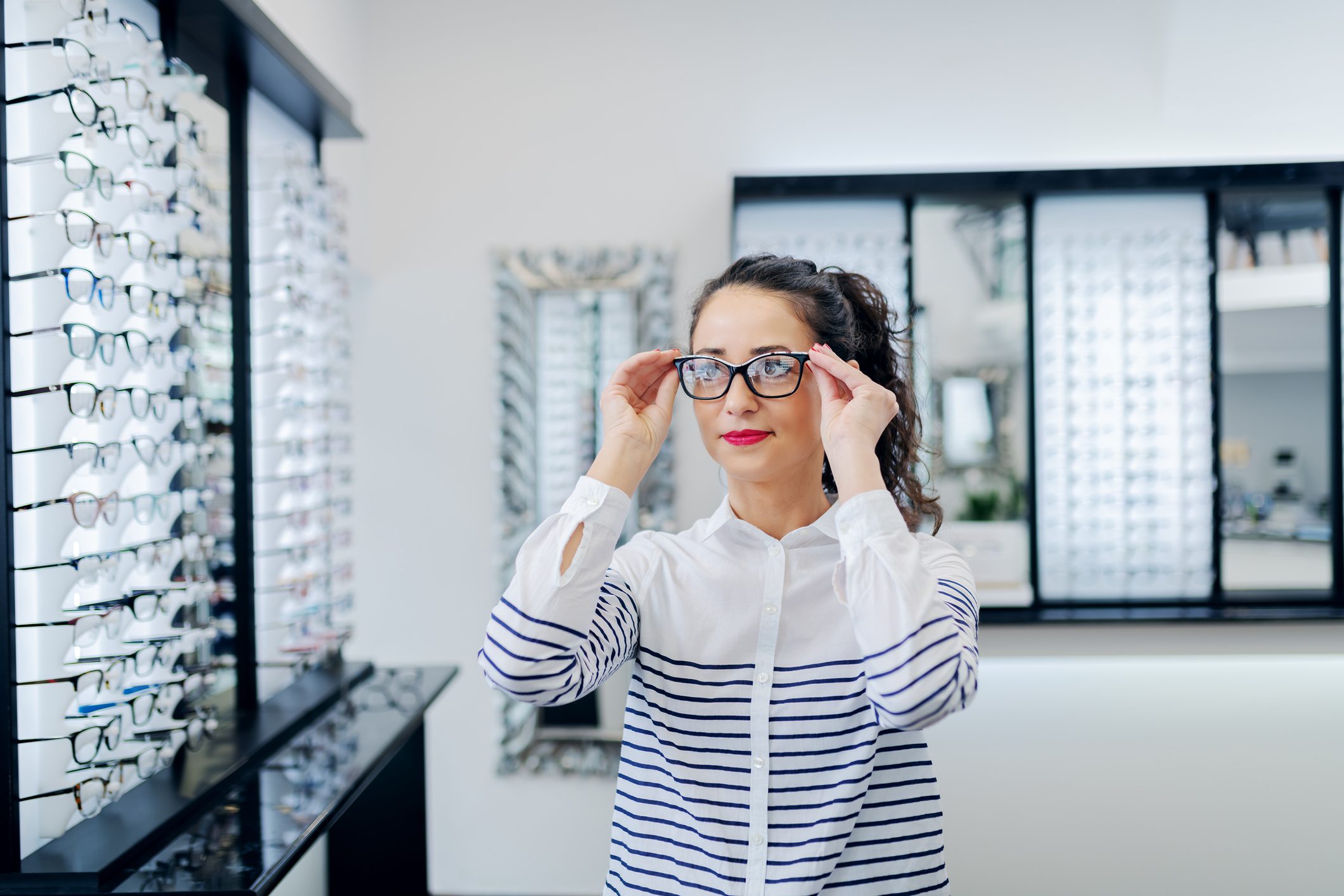 Young brunette woman stands by a row of glasses shelves trying on pairs of glasses in a glasses shop.