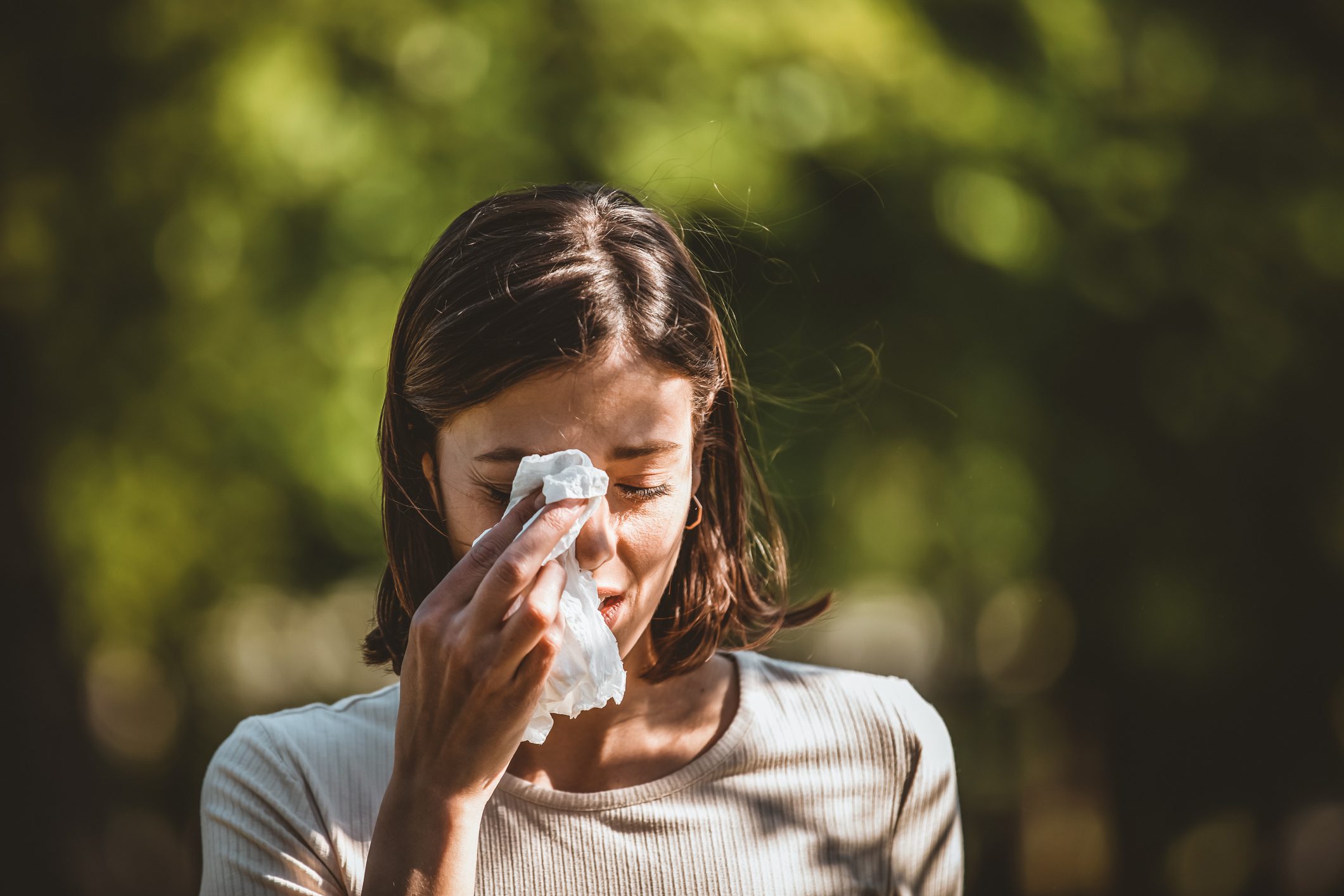 Short-haired brunette woman wipes her eye with a tissue while standing in front of a blurred outdoor background