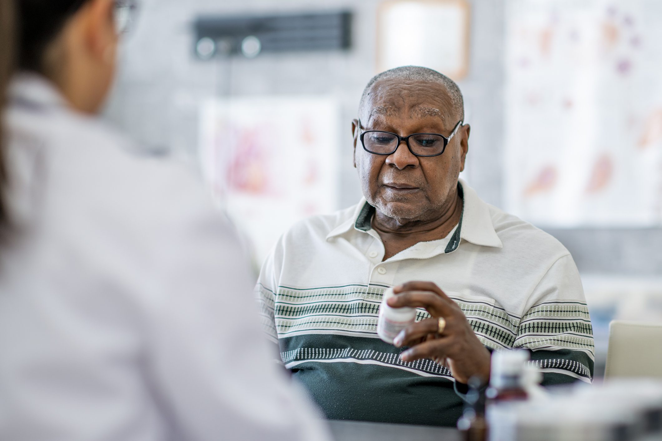 An elderly man in glasses examining a pill bottle with a pharmacist who is obscured in the foreground