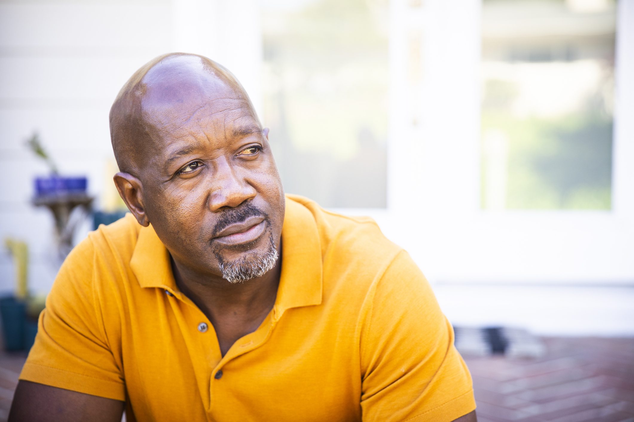 A portrait of an older bald man in a yellow polo sitting outdoors and looking to his left.