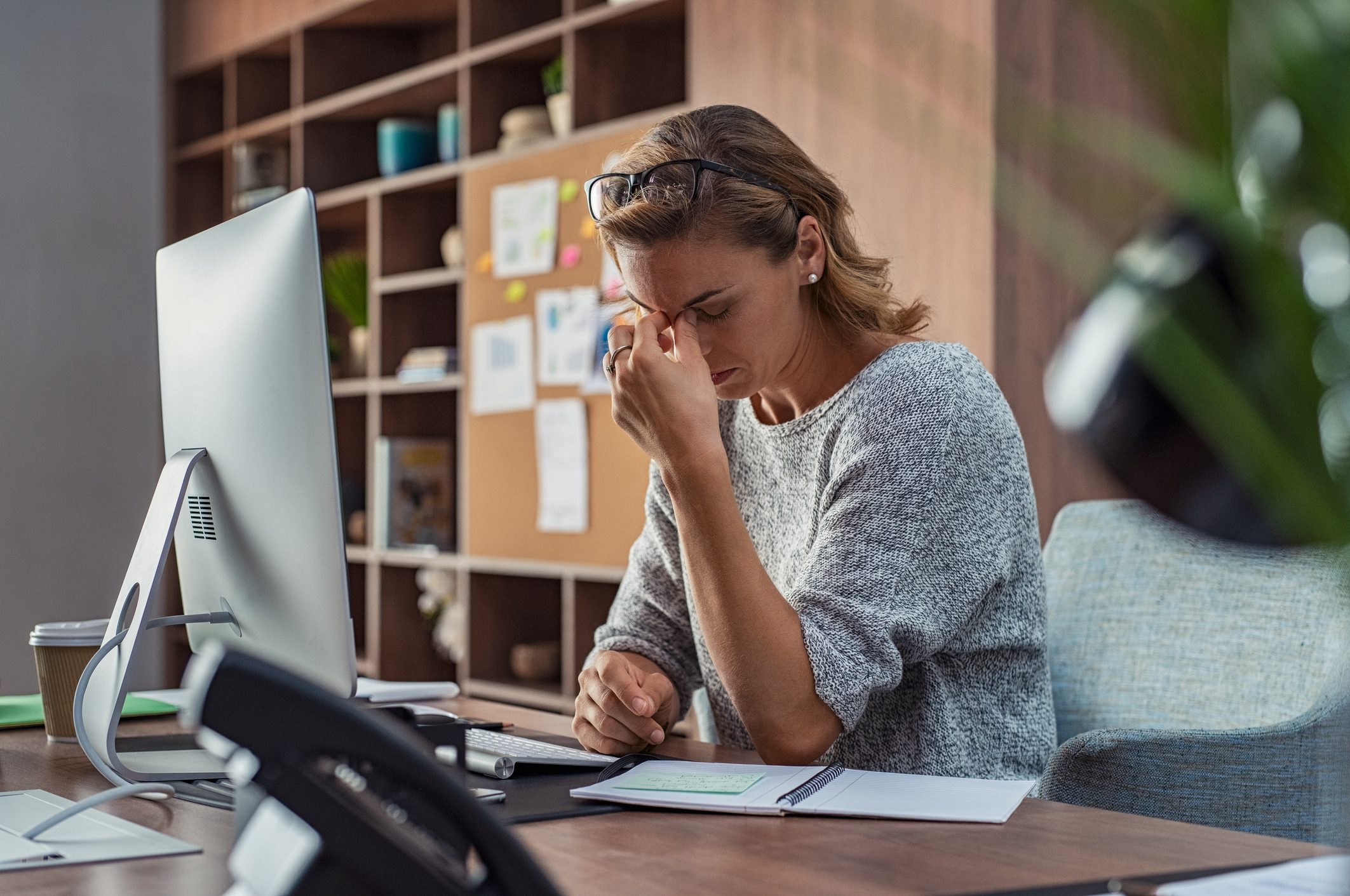 Blonde woman with glasses on her head sits at a desk with a computer and rubs her eyes in a tired way