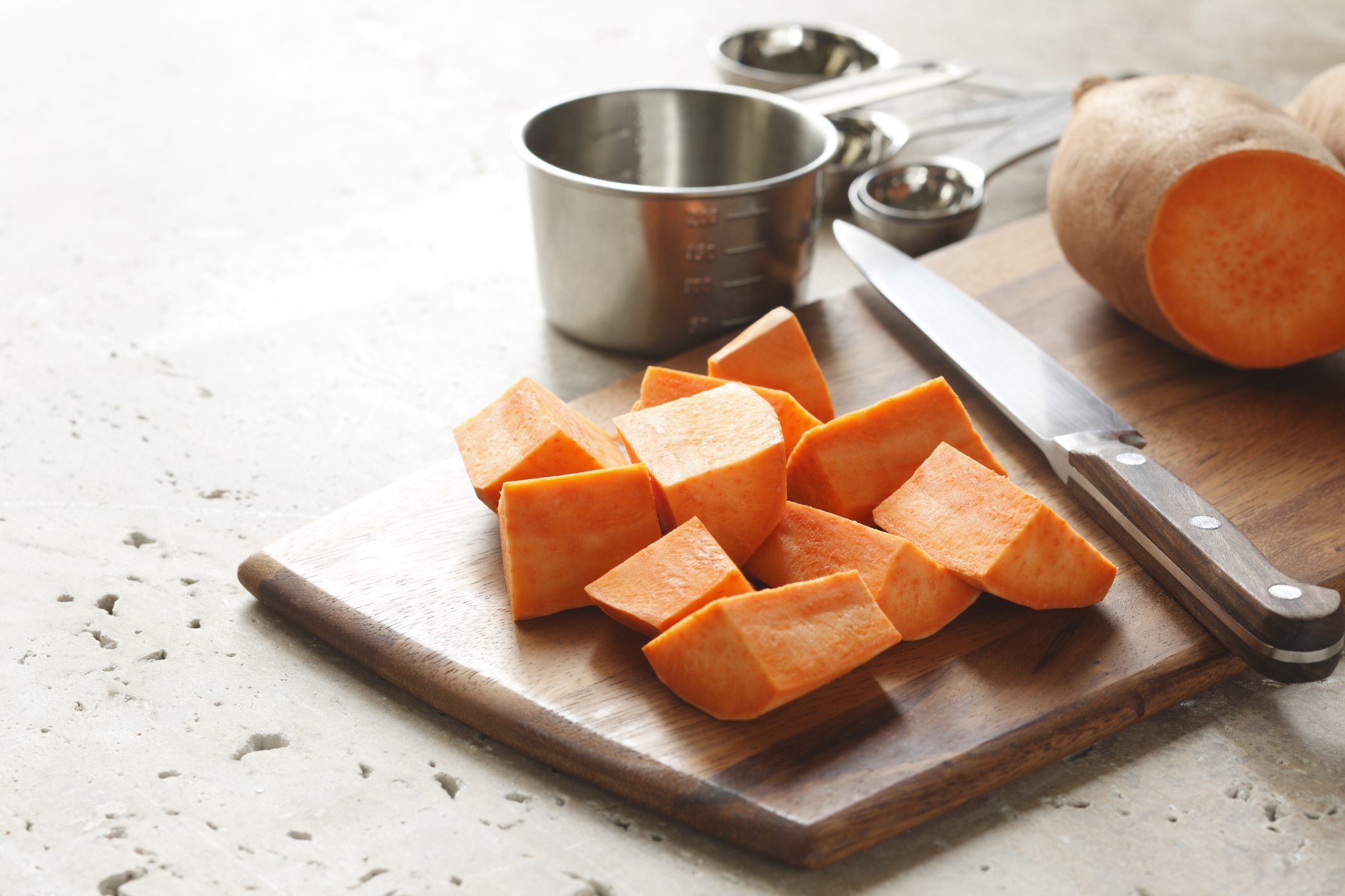 Chopped sweet potato on a cutting board next to a knife and with measuring cups and spoons in the background.