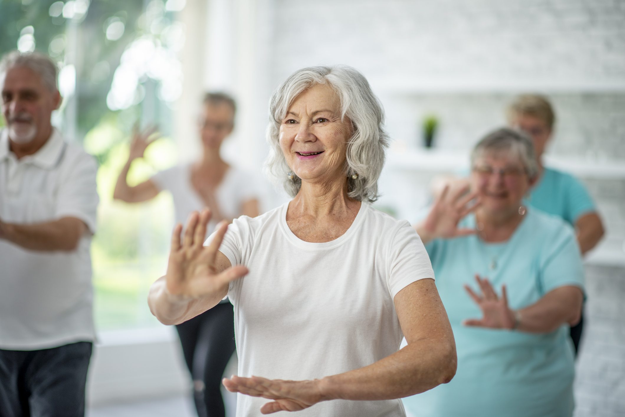 An older woman in a white t-shirt practicing Tai Chi with a group of smiling older adults.