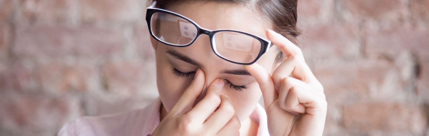 Woman lifts glasses and pinches nose in front of a blurred brick background.