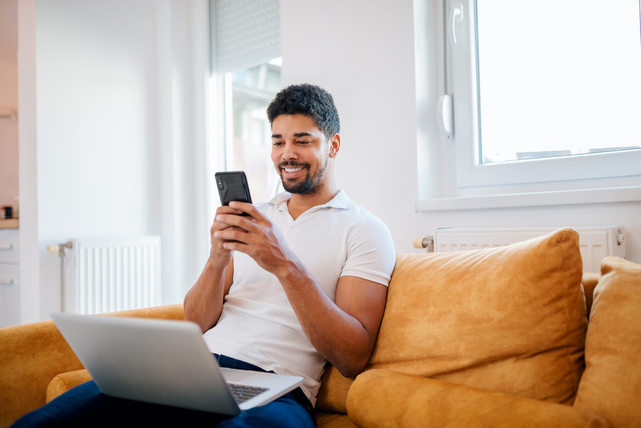 A cheerful man sitting on a sofa with a laptop and checking his phone.