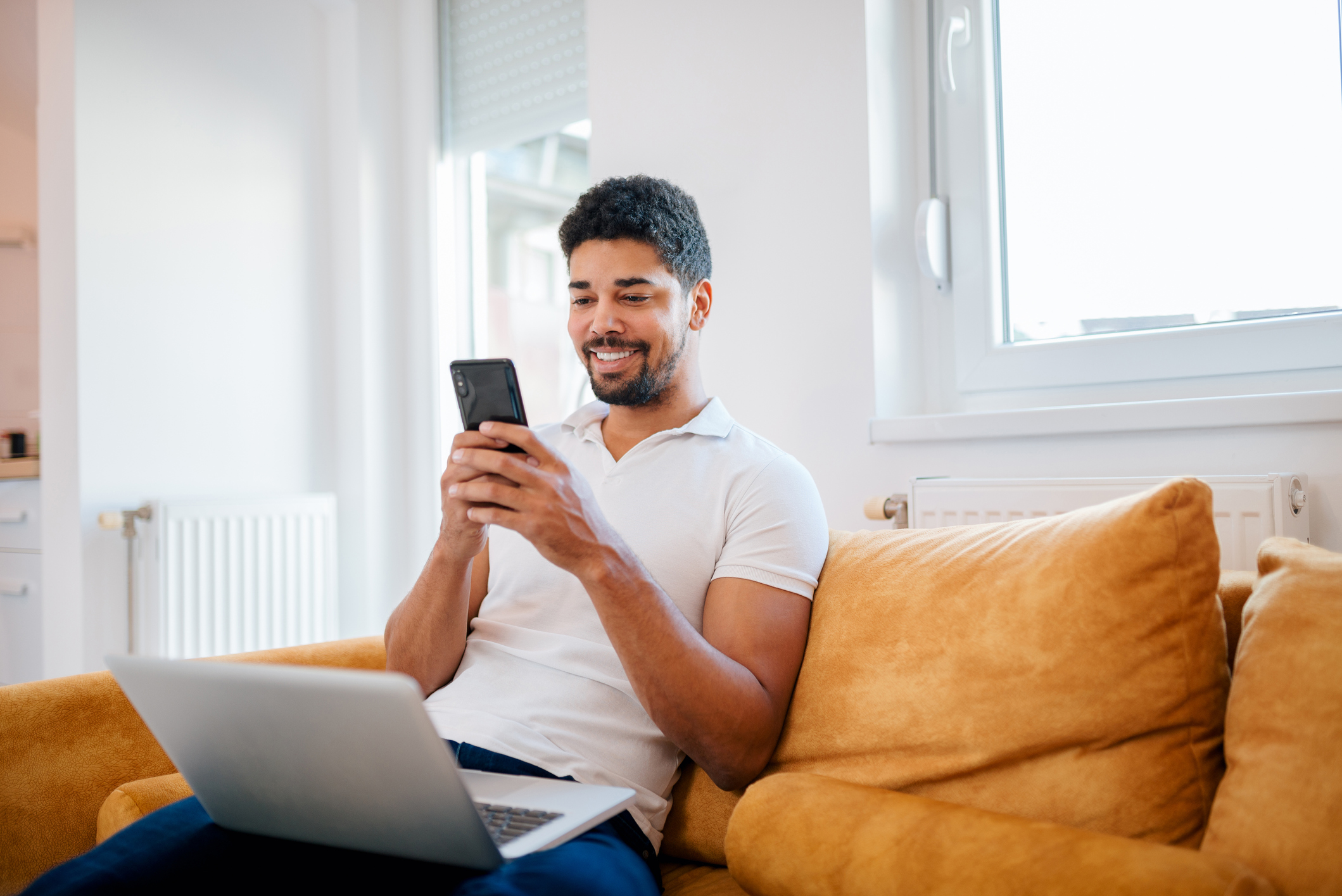 A cheerful man sitting on a sofa with a laptop and checking his phone