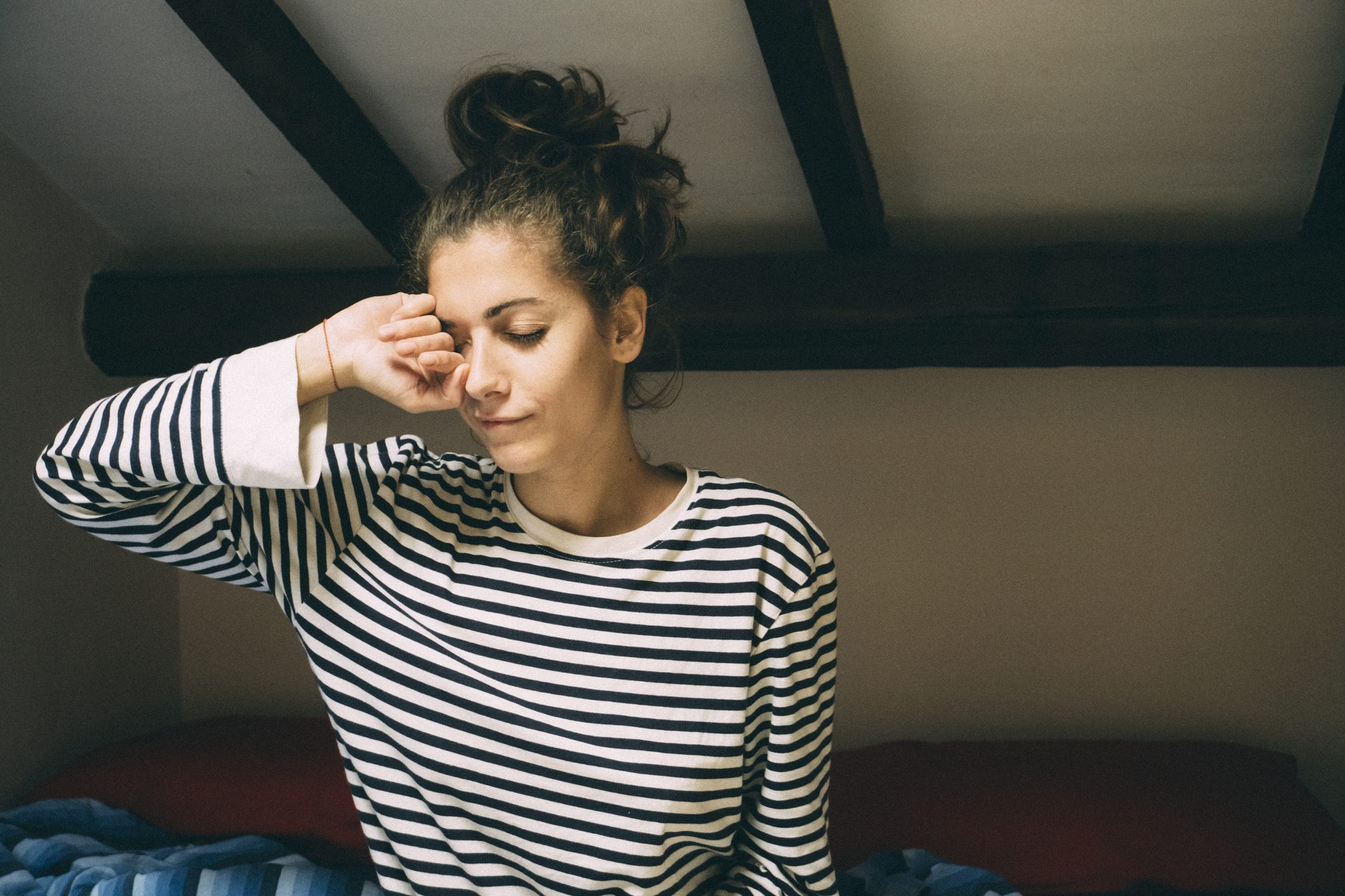 Young woman with brown hair in a striped shirt sits up in bed and rubs her eye