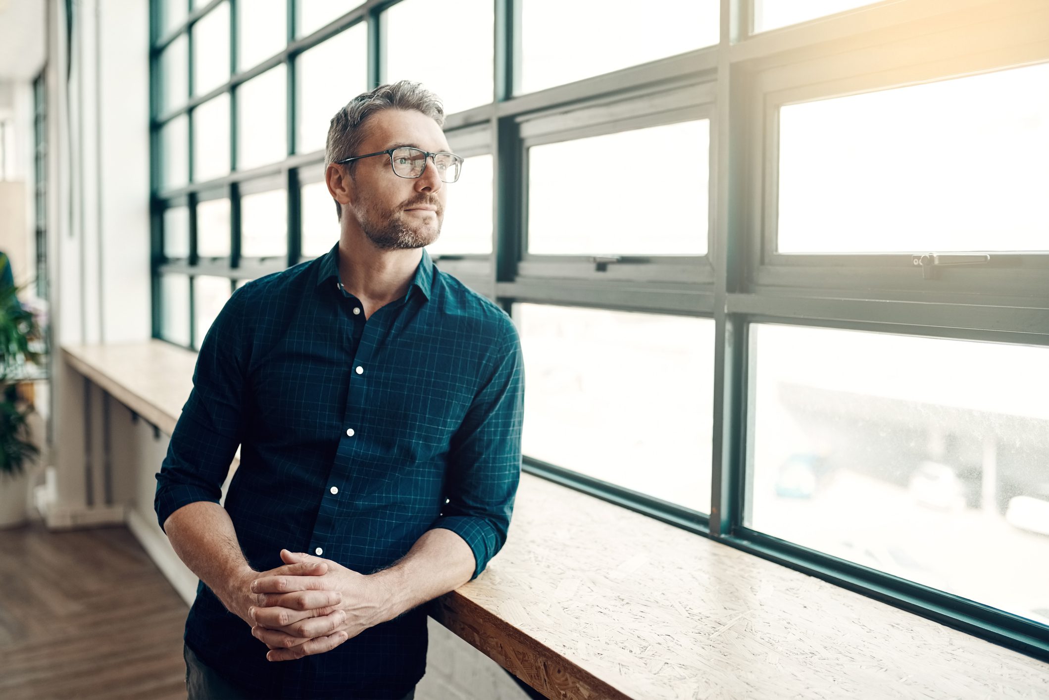 Middle aged man with gray hair, brown scruff, and glasses leans against a wooden counter in front of a long garage-style window