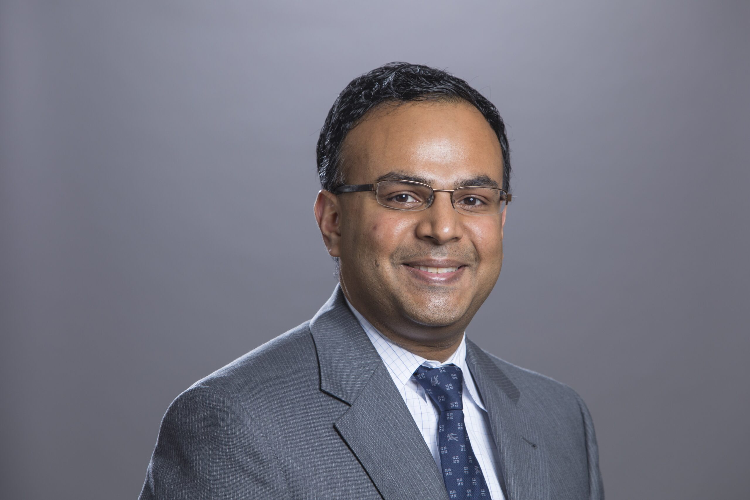 Headshot of a man with glasses, gray blazer, blue tie, and white shirt smiling in front of a gray background