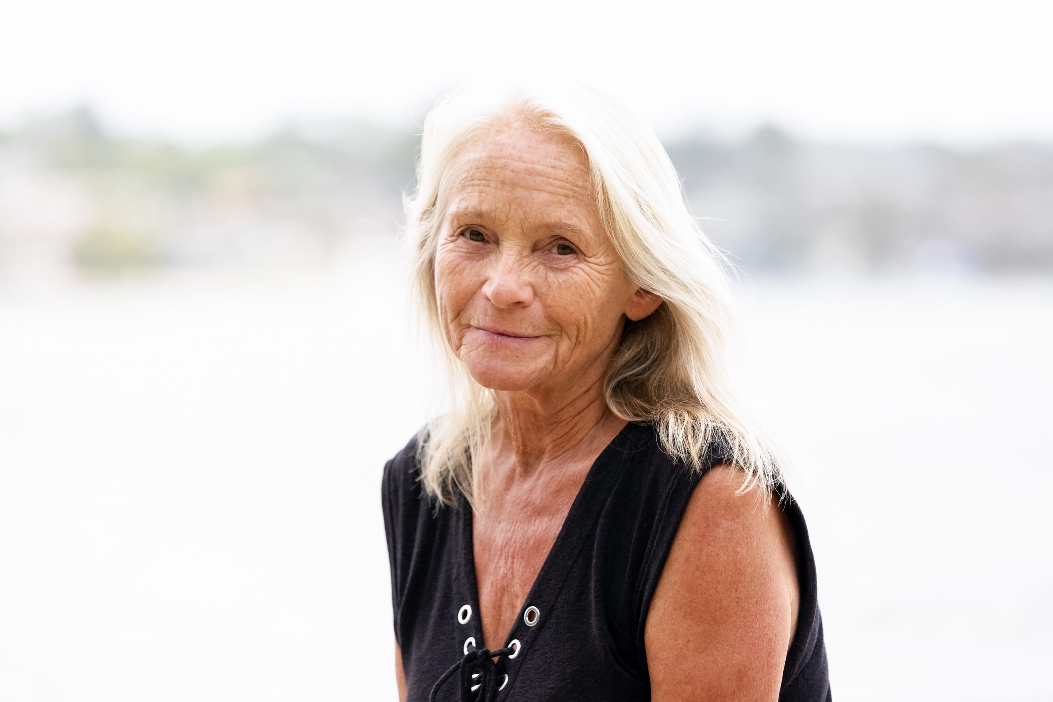 An older woman with white hair in a black tank top sitting in front of a body of water.