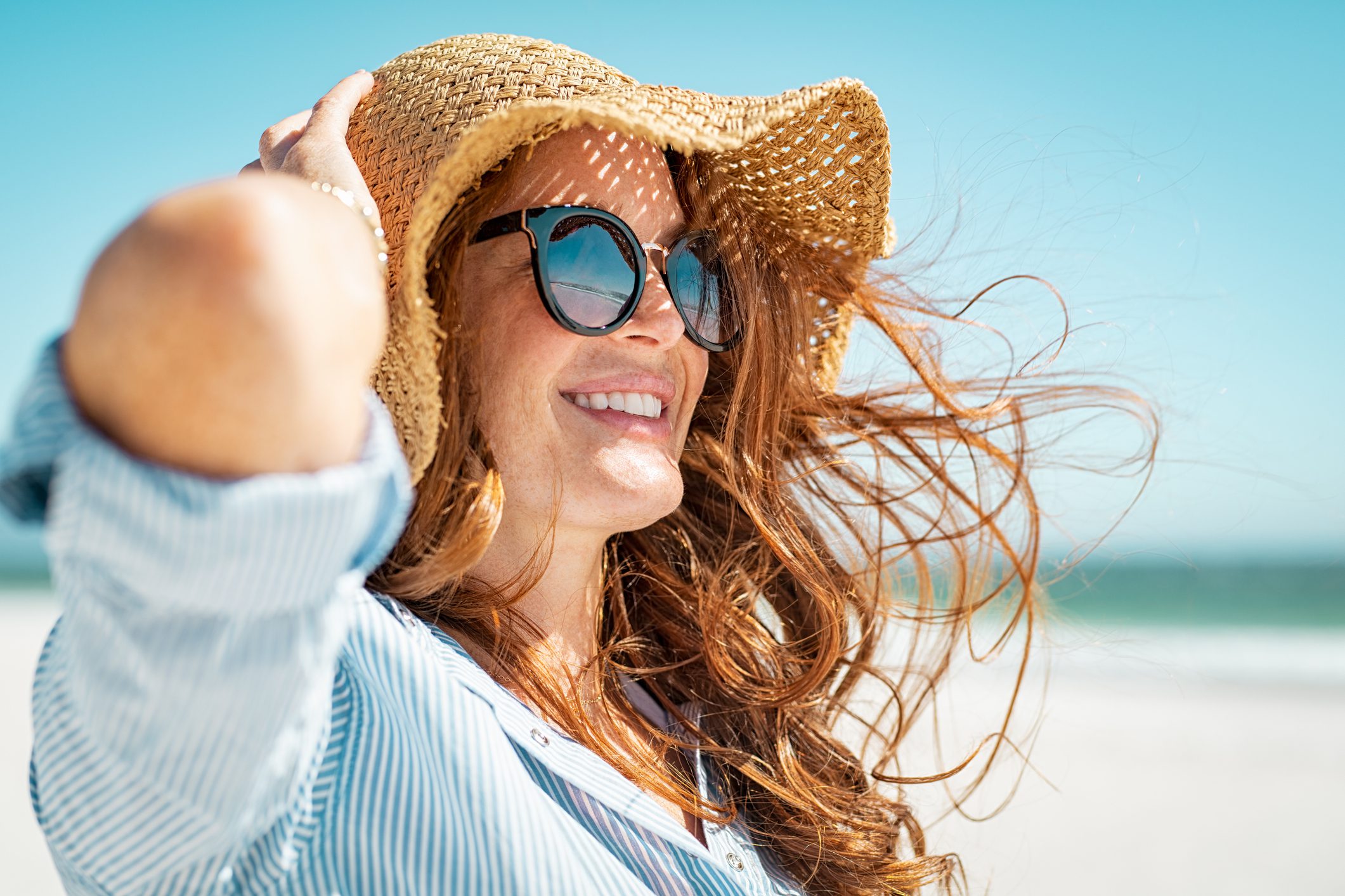 A young, red-haired woman in a hat and sunglasses smiling on the beach and looking at something off camera