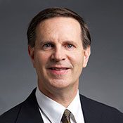 Headshot of a man with brown hair and a suit in front of a gray background