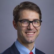 Headshot of a man with glasses smiling at the camera in front of a gray background
