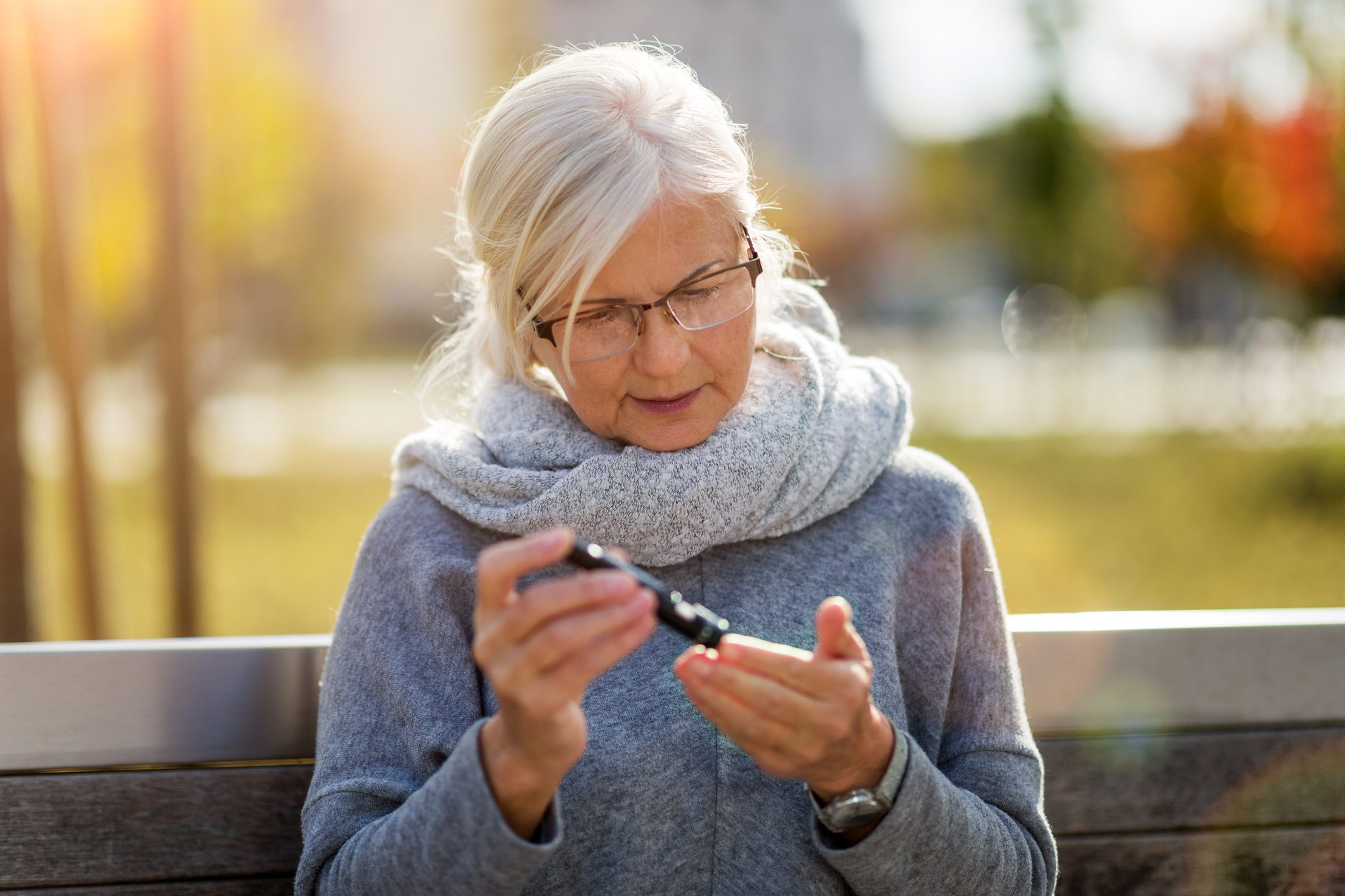 Older woman with white hair, glasses, and a gray scarf and sweater sits on a bench and performs a finger prick test on herself.