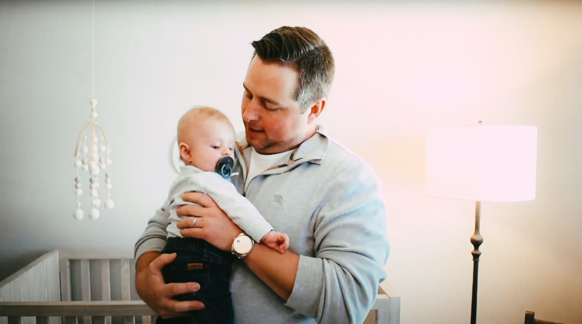 Brown-haired man holding a baby with a pacifier in a home nursery.