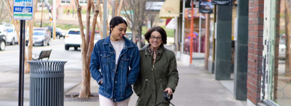 Young woman in a denim jacket walks down a city street with brunette woman in an olive green jacket.