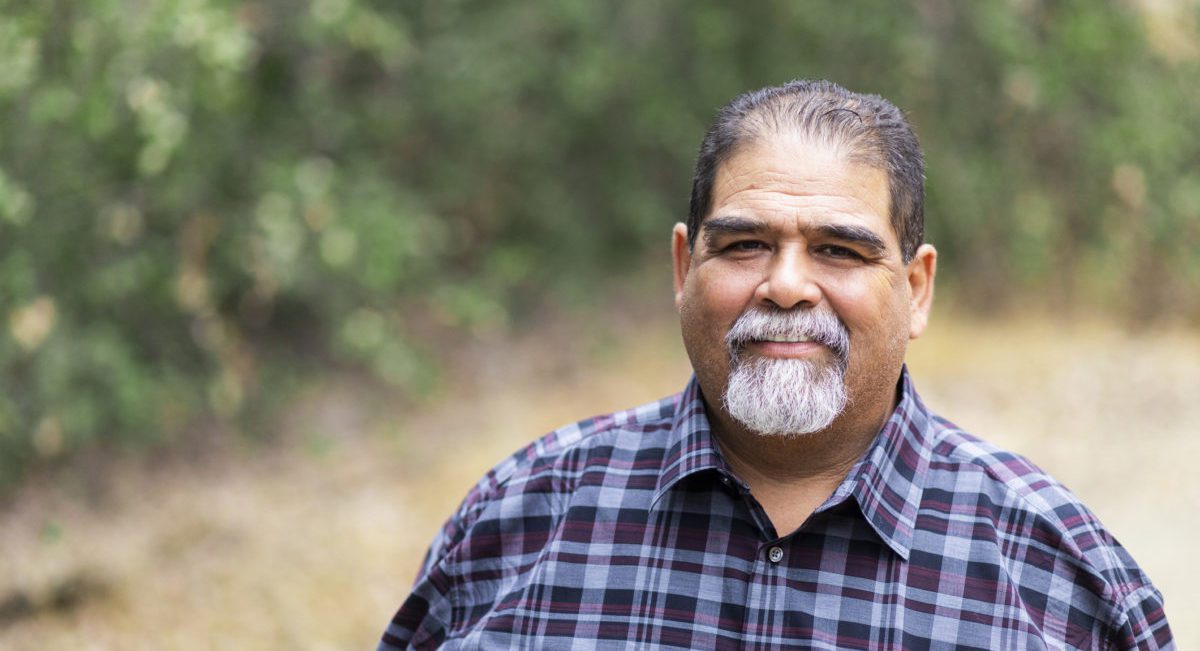 Older man with a white beard and mustache and black hair smiles in front of a blurred forest background
