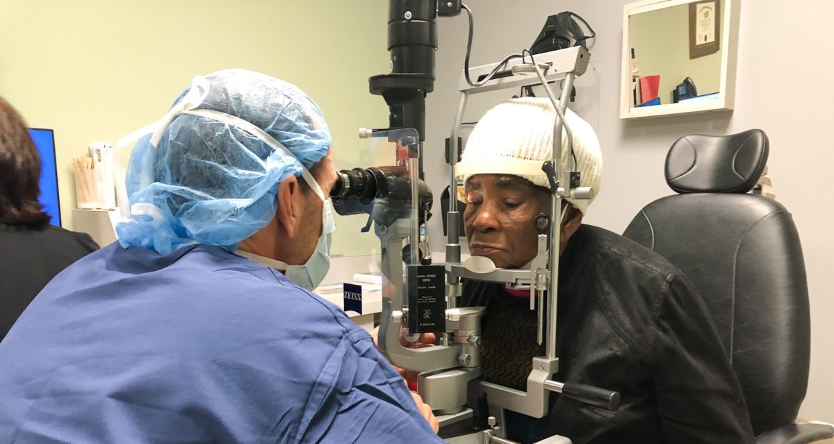 An eye doctor in scrubs examining a patient in a white hat using a microscope