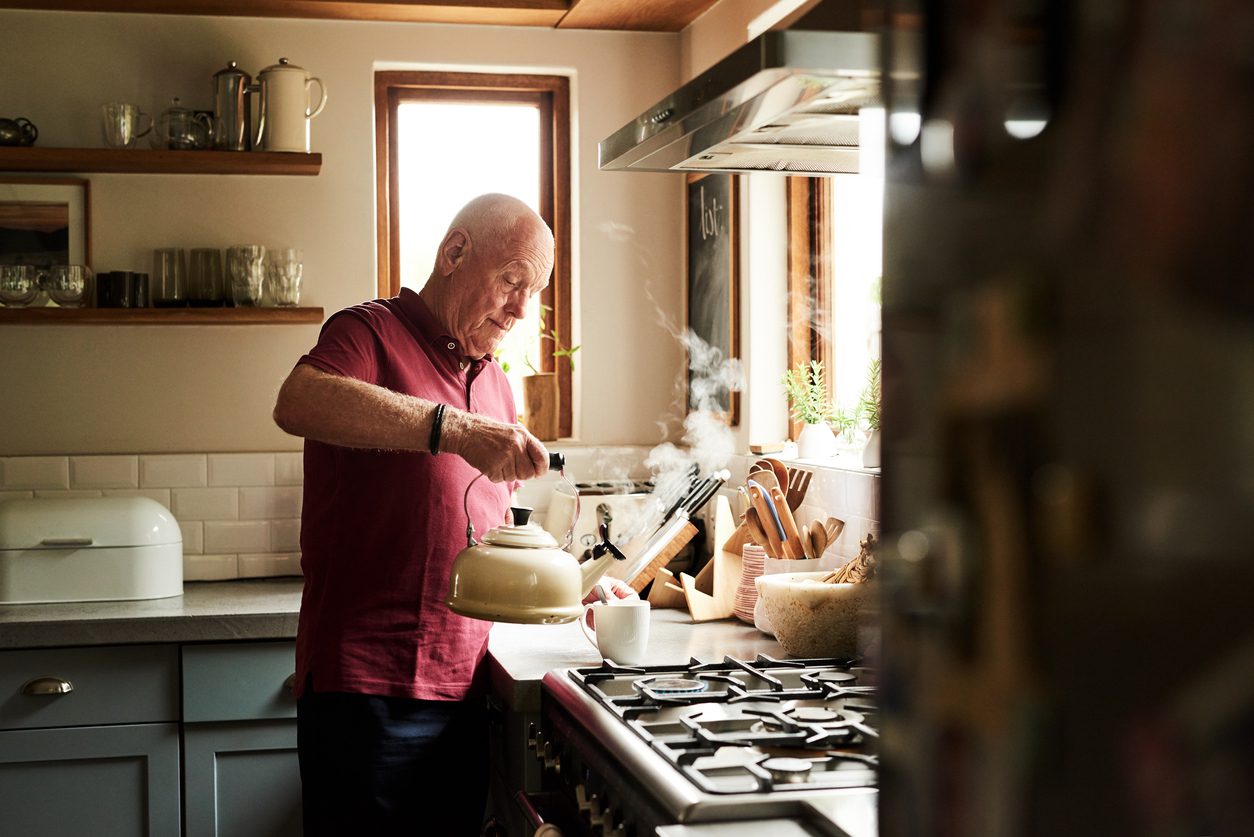 In a kitchen, a bald older man in a red polo removes a steaming kettle from the stovetop and prepares to pour its contents into a mug