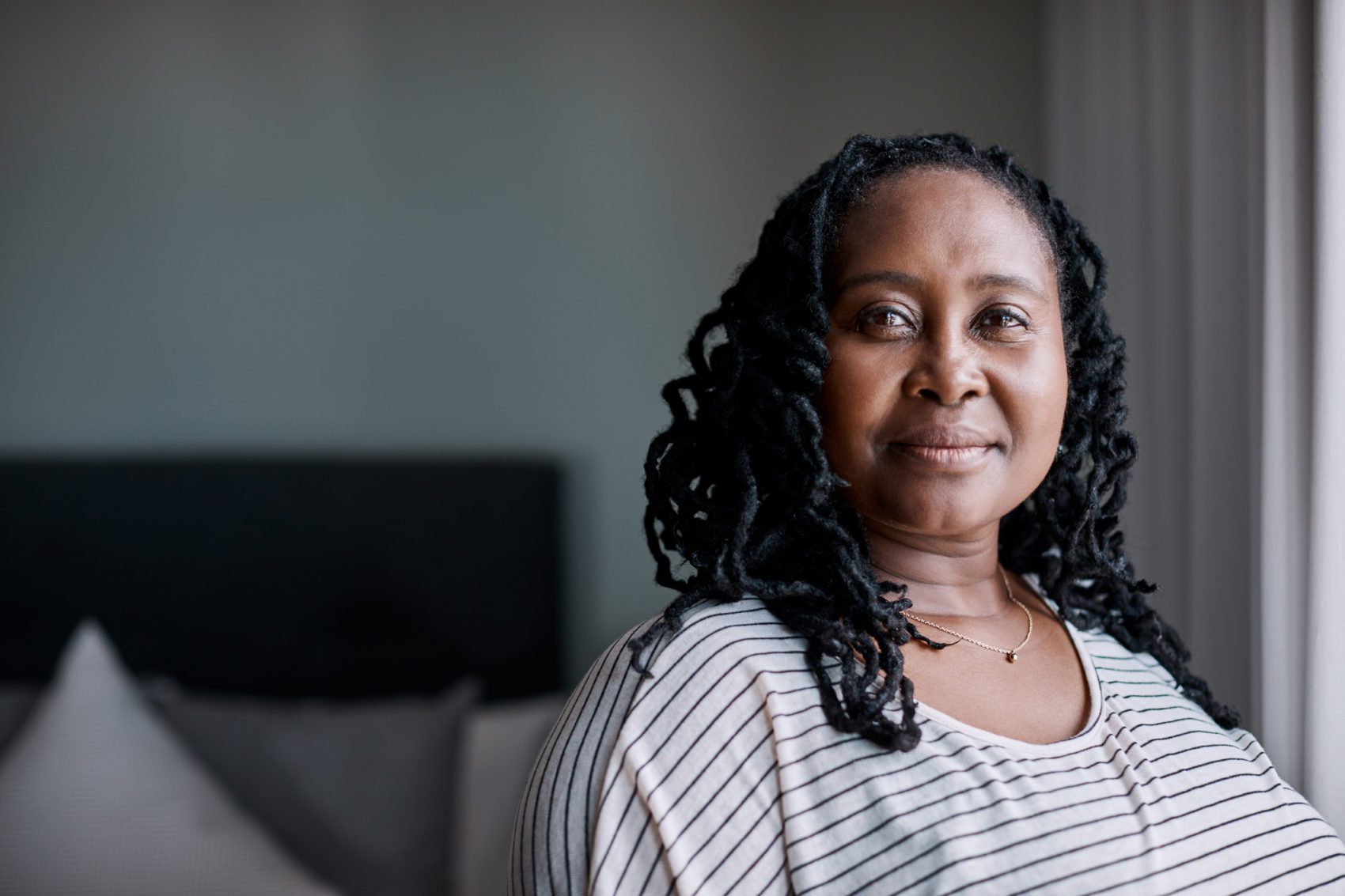 Black woman with curly black hair and white striped shirt soft smiles at the camera in front of a blurred bedroom background