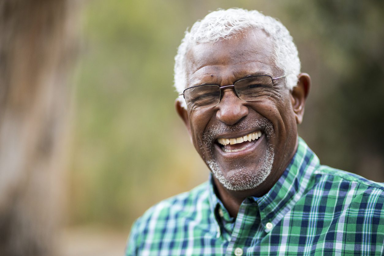 Older man with glasses and gray scruff laughs while wearing a green and blue plaid shirt