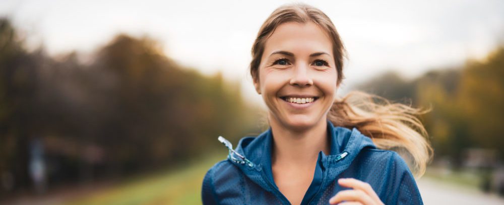 Blonde woman with a ponytail and blue workout jacket smiles as she runs on an autumn day.