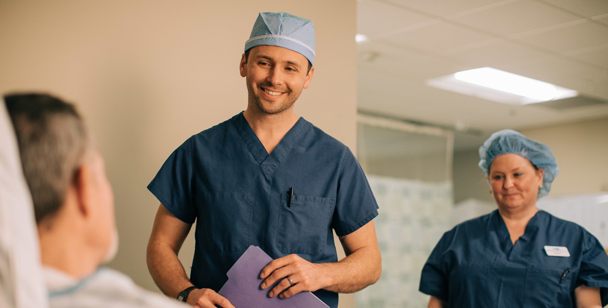 Young male doctor in dark blue scrubs and scrub cap and similarly-attired female staffer smile at a male patient in a bed