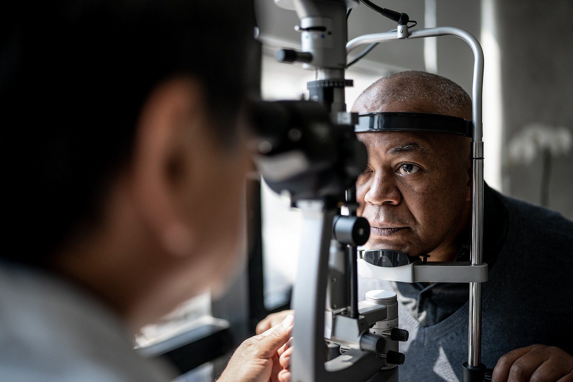 Older male patient puts chin on chin rest during an eye exam with a doctor who is obscured in foreground