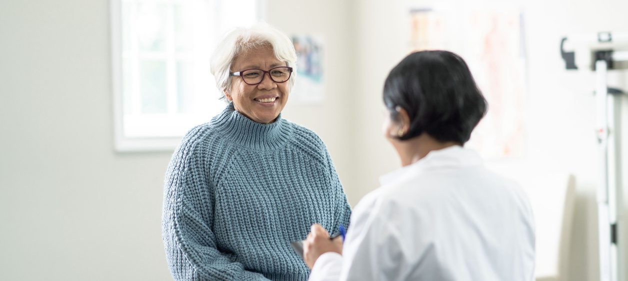 Older female patient with white hair, glasses, and blue sweater talks to female doctor who is facing away from the camera