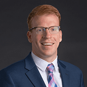 Headshot of a red haired man in glasses, a blue blazer, and a blue and pink tie in front of a dark background