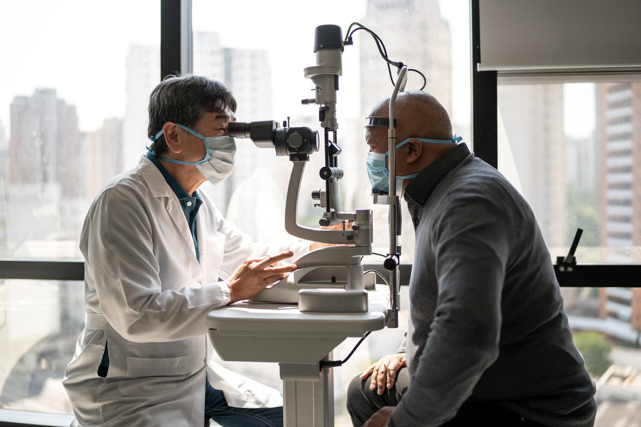 Male doctor in white coat and mask examines a bald male patient in a mask in an exam room with a large window