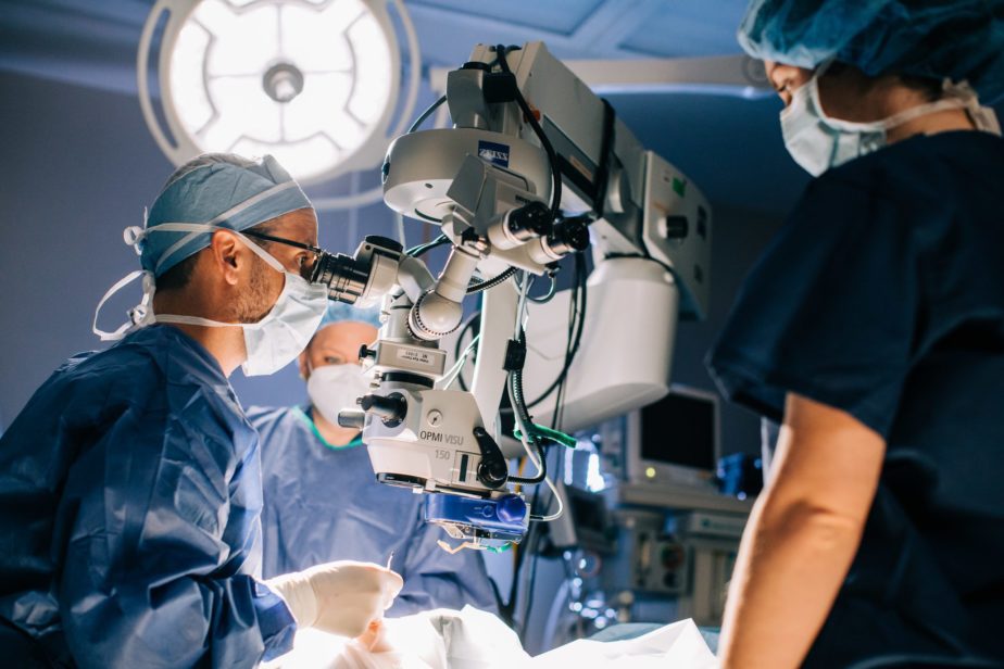 Male doctor in scrubs and mask uses a surgical microscope in the operating room while assisted by other doctors