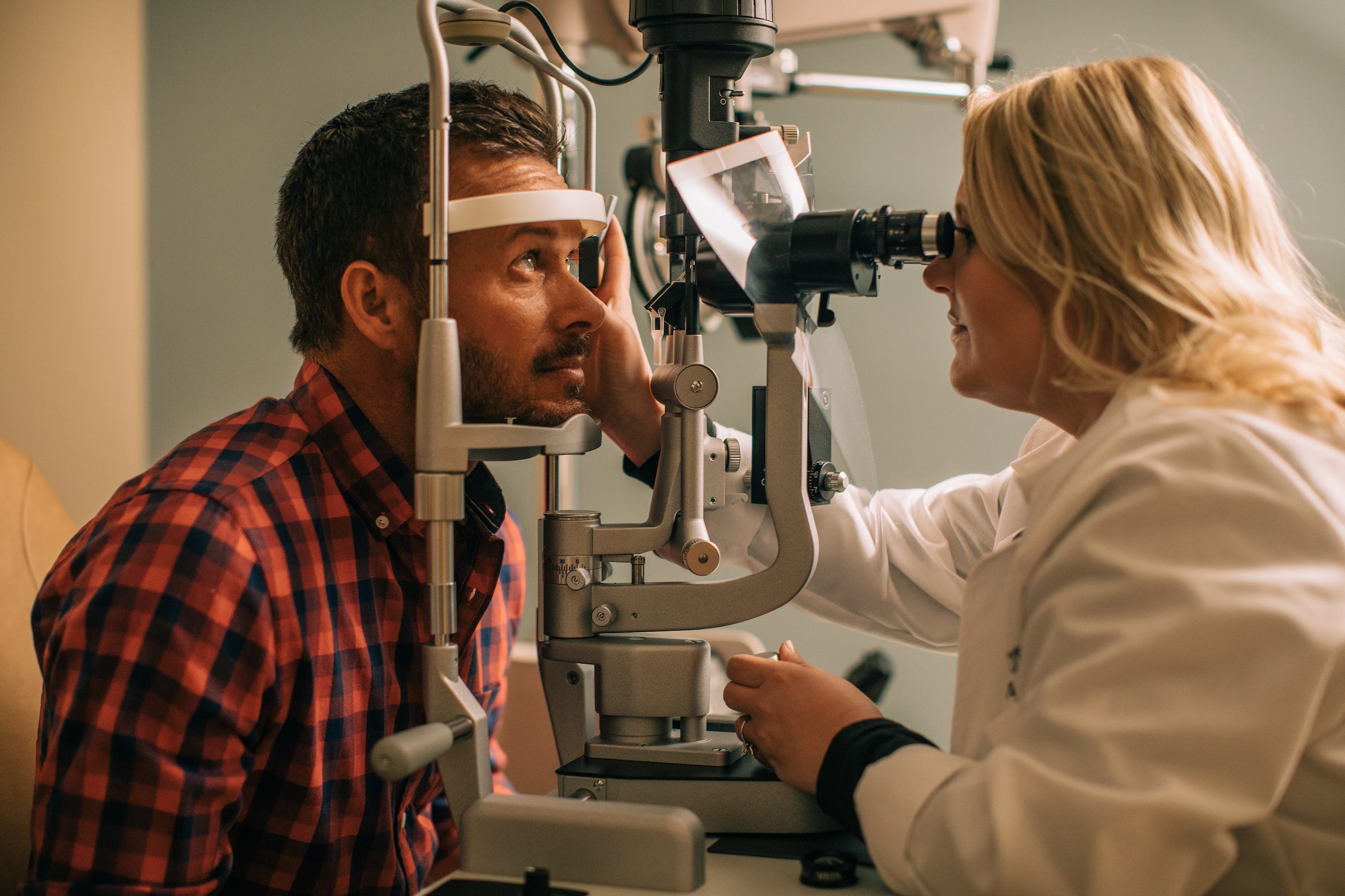 Blonde female doctor in a white coat examines the eyes of a dark-haired male patient in a red and blue flannel