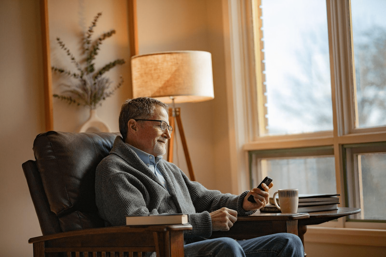 Older man with glasses and a gray sweater relaxes in a leather chair with a mug of coffee and smiles at his smartphone