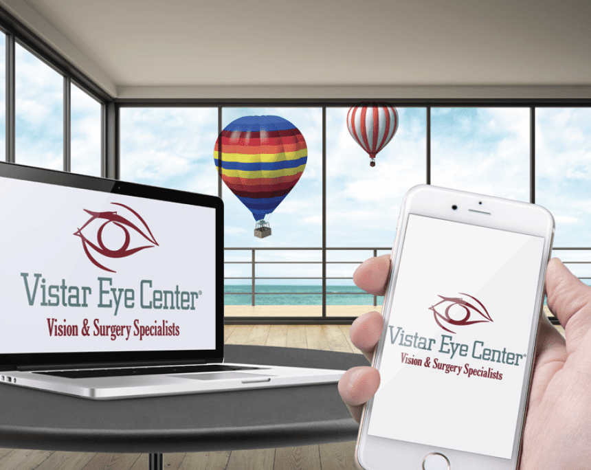 Close-up of a laptop and smartphone displaying the Vistar Eye Center logo, with the tagline 'Vision & Surgery Specialists.' In the background, large windows reveal a scenic view of hot air balloons floating over a body of water.